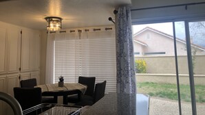Un comedor con mesa y sillas, un lámpara en el techo y vista a una casa a través de una ventana con persianas.