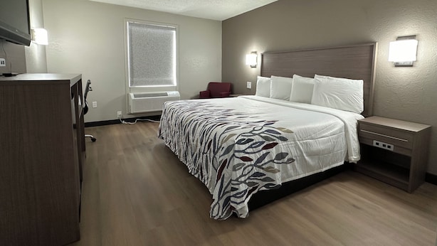 A hotel room with a bed, bedside table, lamp, and a window with blinds. - Red Roof Inn Richmond, VA – W Broad St (Richmond)