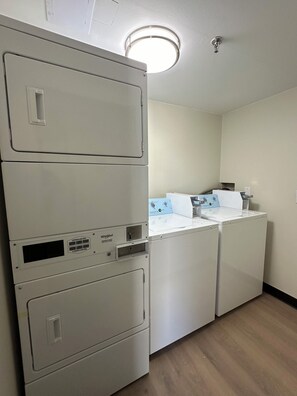 A laundry room with stacked washer and dryer units, a control panel, and a ceiling light. - Red Roof Inn Richmond, VA – W Broad St (Richmond)