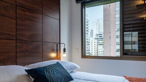 A modern hotel room with a large bed, a wooden headboard, a window with a view of tall buildings, and a bedside lamp. - Haddock_166' 1 bedroom apartment in Jardins (São Paulo)