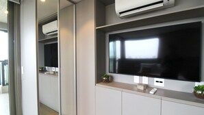 A modern bathroom with a large mirror, a wall-mounted air conditioner, a flat-screen TV, and a small plant on the vanity.