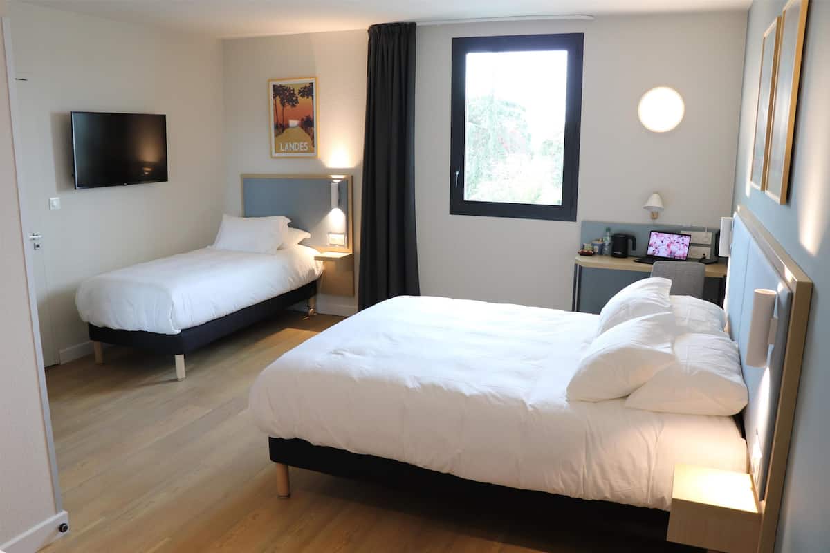 A hotel room with two beds, a TV, a desk with a computer, and a window with a view of trees.