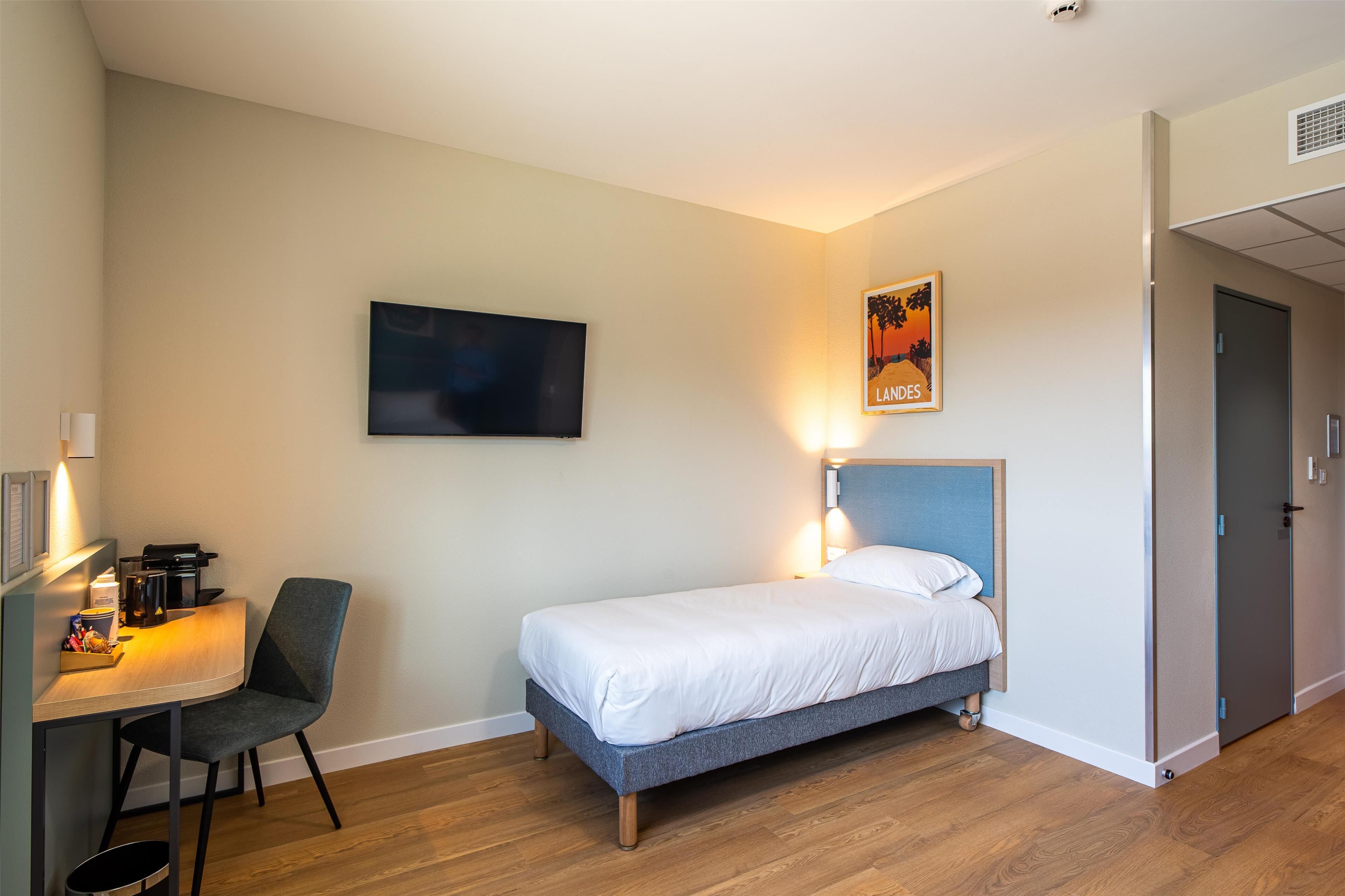 Ett hotellrum med en säng, ett skrivbord med en stol, en platt-TV och en inramad tavla på väggen.