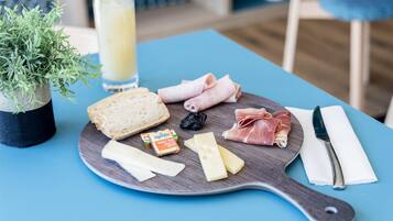 Un tagliere con diversi formaggi, salumi e pane su un tavolo di legno, accompagnato da un bicchiere di succo di frutta e una piccola pianta in vaso.