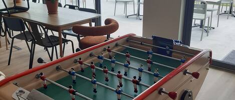 A foosball table in a room with large windows, potted plants, and outdoor seating visible through the windows.