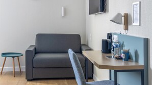Una habitación moderna con un sofá gris, una mesita redonda pequeña, un escritorio con una silla, un televisor montado en la pared y una lámpara de pared.