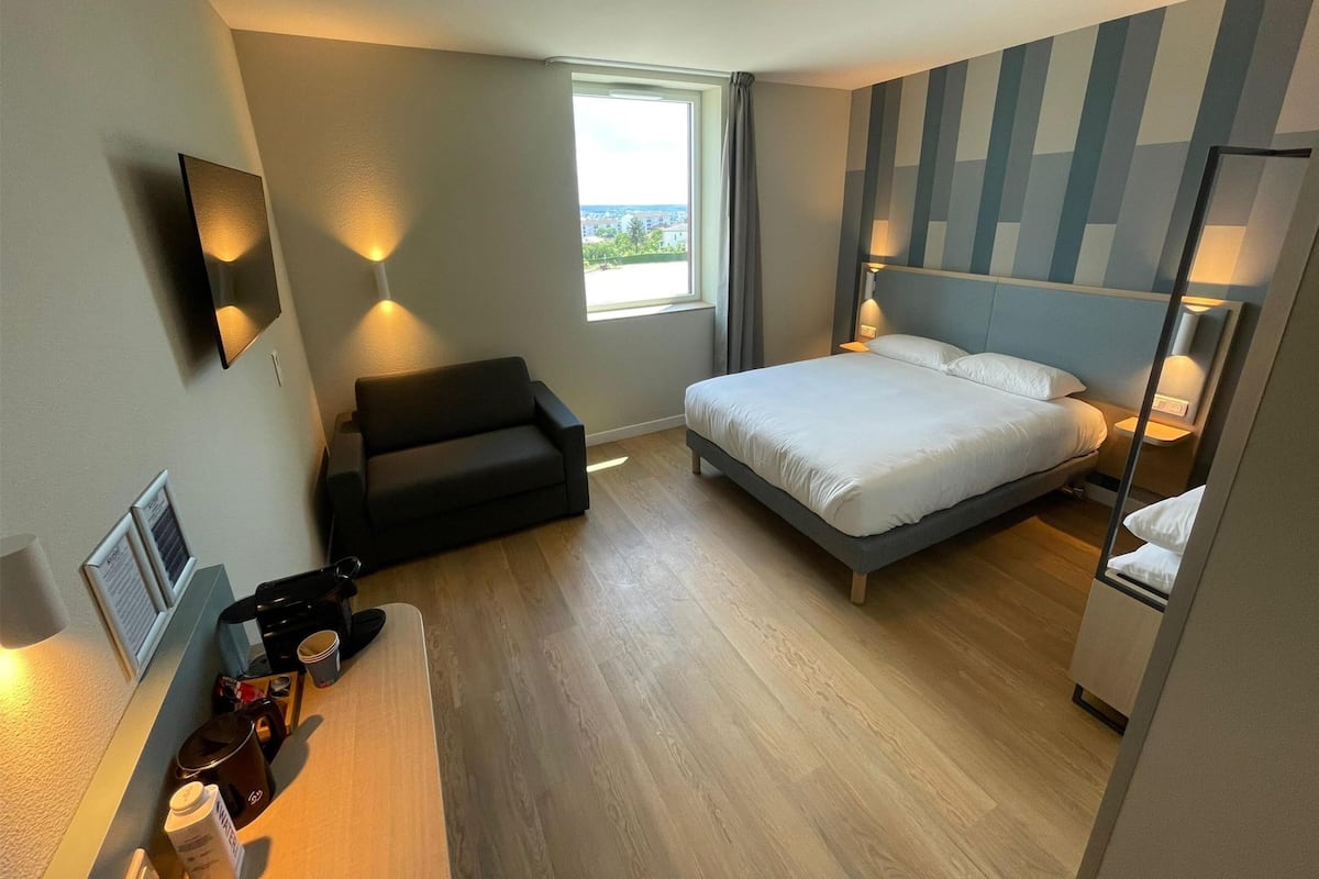 A modern hotel room with a bed, a sofa, a desk with a coffee maker, and a window with a view.