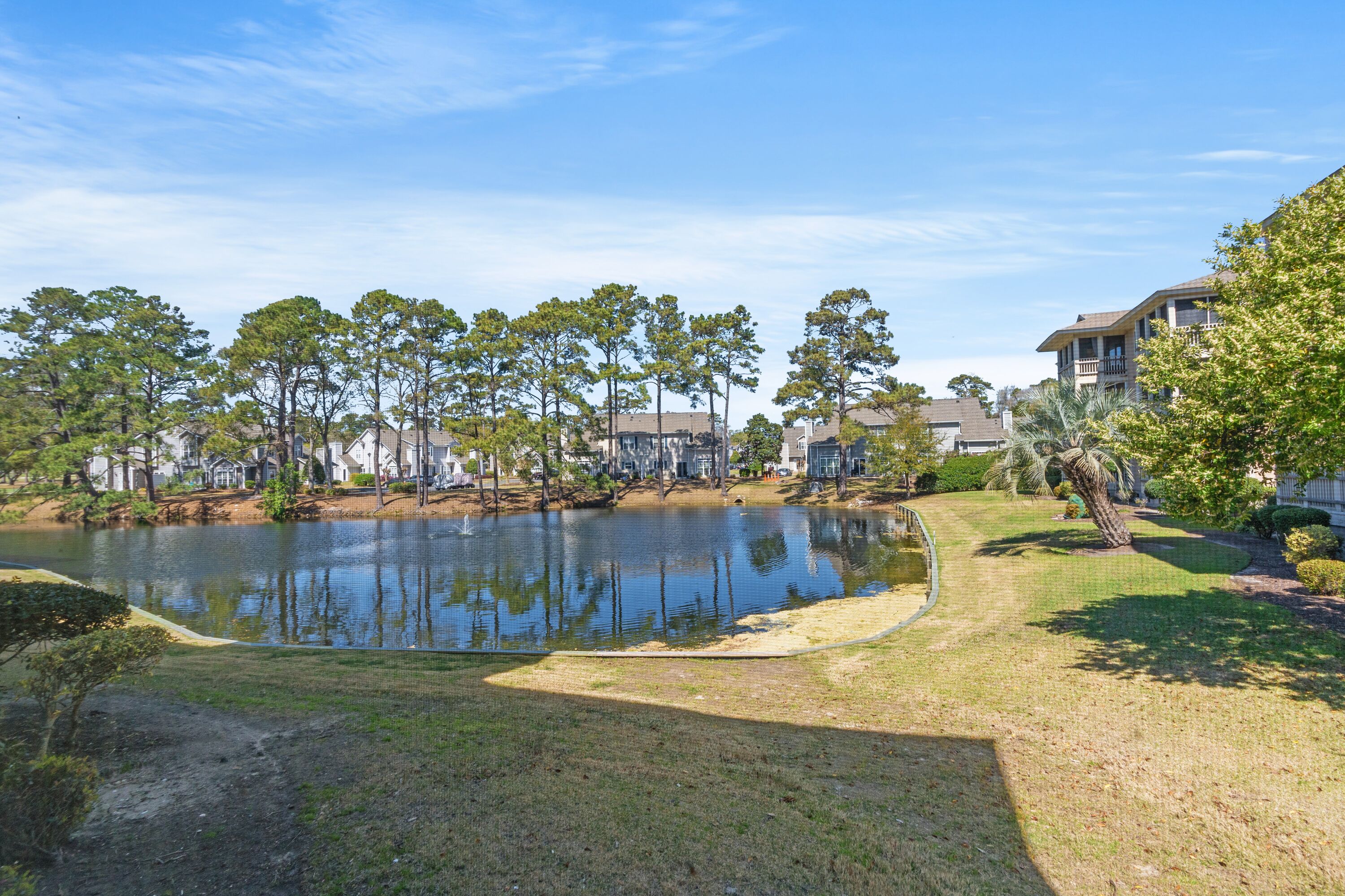 A serene lakeside view with a well-maintained lawn, trees, and residential buildings in the background.