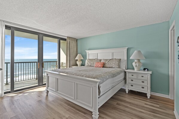 A bedroom with a large bed, a nightstand, a lamp, and a view of the ocean through a sliding door.