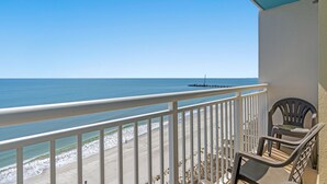 A balcony with a view of the beach and ocean. There are two chairs and a table on the balcony.