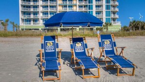 Three beach chairs under a blue umbrella with a North Myrtle Beach logo, in front of a multi-story hotel with balconies.