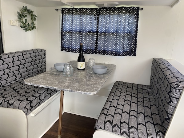 Una kitchenette compacta con una mesa, dos sillas, una botella de vino y una ventana con cortinas estampadas.