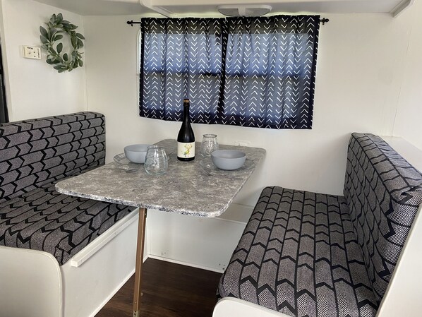 A compact kitchenette with a table, two chairs, a wine bottle, and a window with patterned curtains.