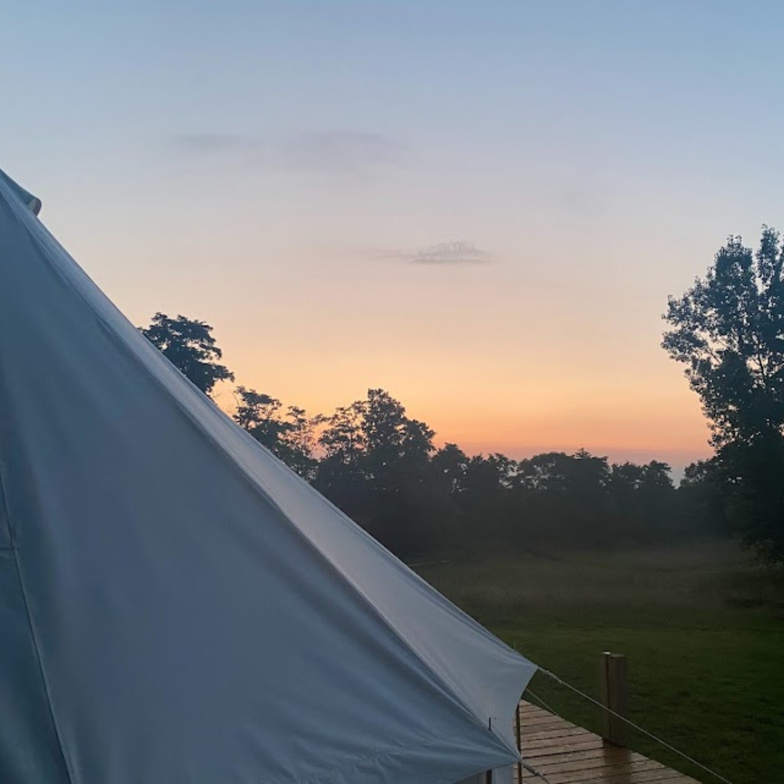 Une tente dotée d’une terrasse en bois, installée dans un cadre verdoyant, avec des arbres en arrière-plan et un ciel dégagé.