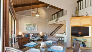 A spacious living area with a dining table set for four, surrounded by wicker chairs. A staircase with wooden railings leads to an upper level.