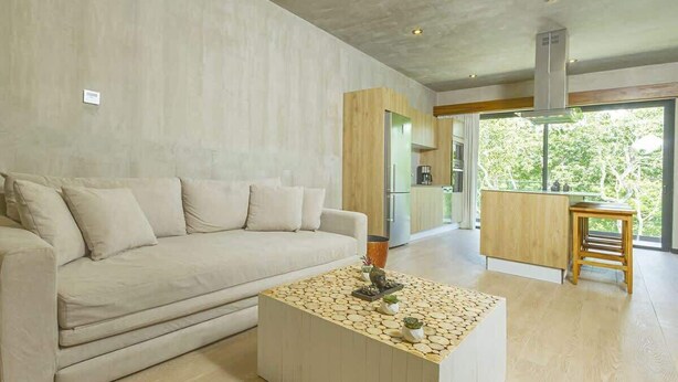 A modern living room with a beige sofa, a wooden coffee table with plants, and a kitchen area visible through a glass door. - Relaxing 1 BDRM, 1 BR, Balcony & Pool Access Apt (Tulum)
