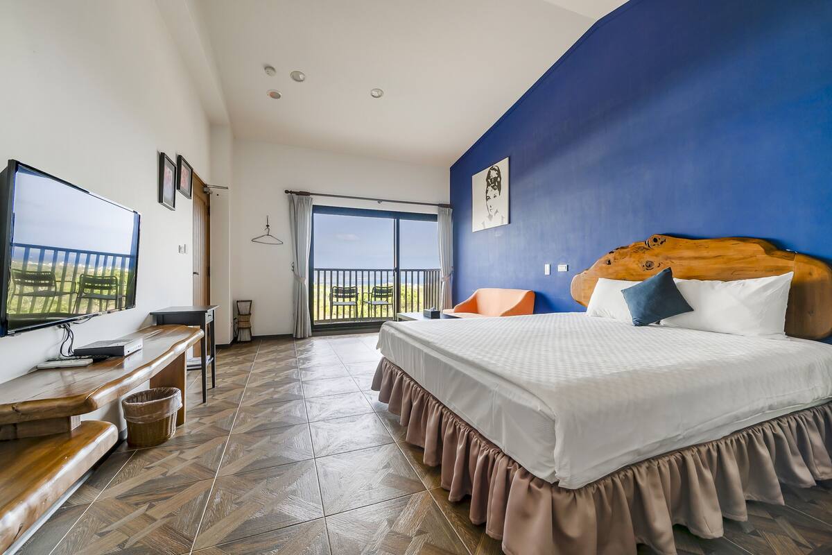 A bedroom with a large bed, a TV, a bench, and a balcony with a view.
