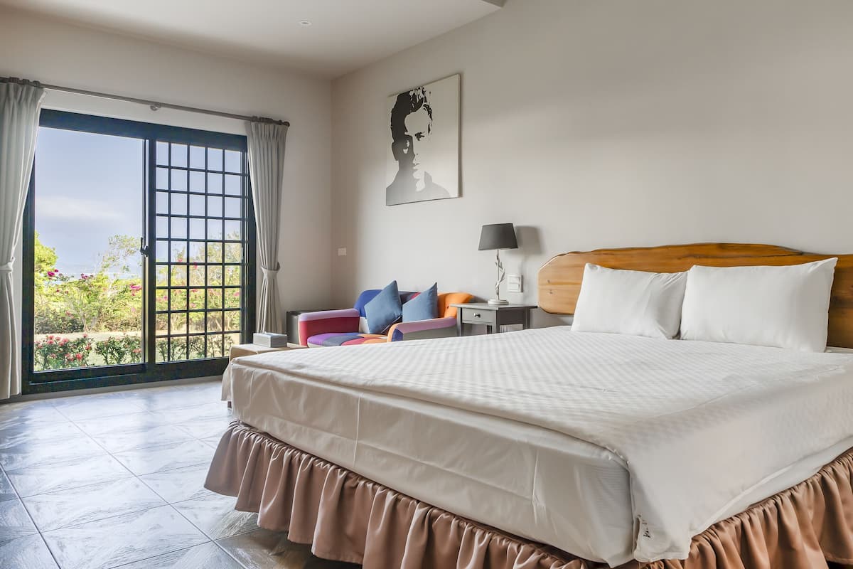 A bedroom with a large bed, a chair, a nightstand, and a window with curtains.