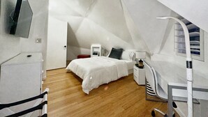 Un dormitorio con techo inclinado, una cama, una mesita de noche, una lámpara, una silla y un televisor colocado en la pared.