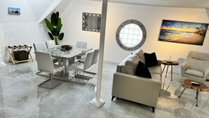 Una sala de estar moderna con piso de mármol, un comedor con mesa de vidrio, un sofá y un sillón. Se ve una ventana grande con vista a la playa y un cuadro en la pared.