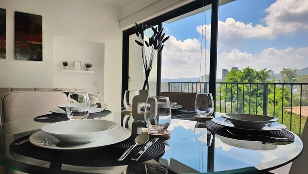 A dining table set with plates, glasses, and cutlery, with a view of greenery and buildings through large windows. - PanoramaTWR 2Brm Short walk to everything Pool&Gym

 (San Salvador)