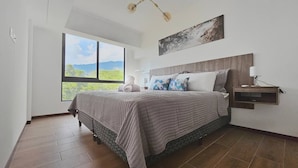A bedroom with a large window, a bed with a quilted bedspread, a nightstand with a lamp, and a wall-mounted shelf. - PanoramaTWR 2Brm Short walk to everything Pool&Gym

 (San Salvador)