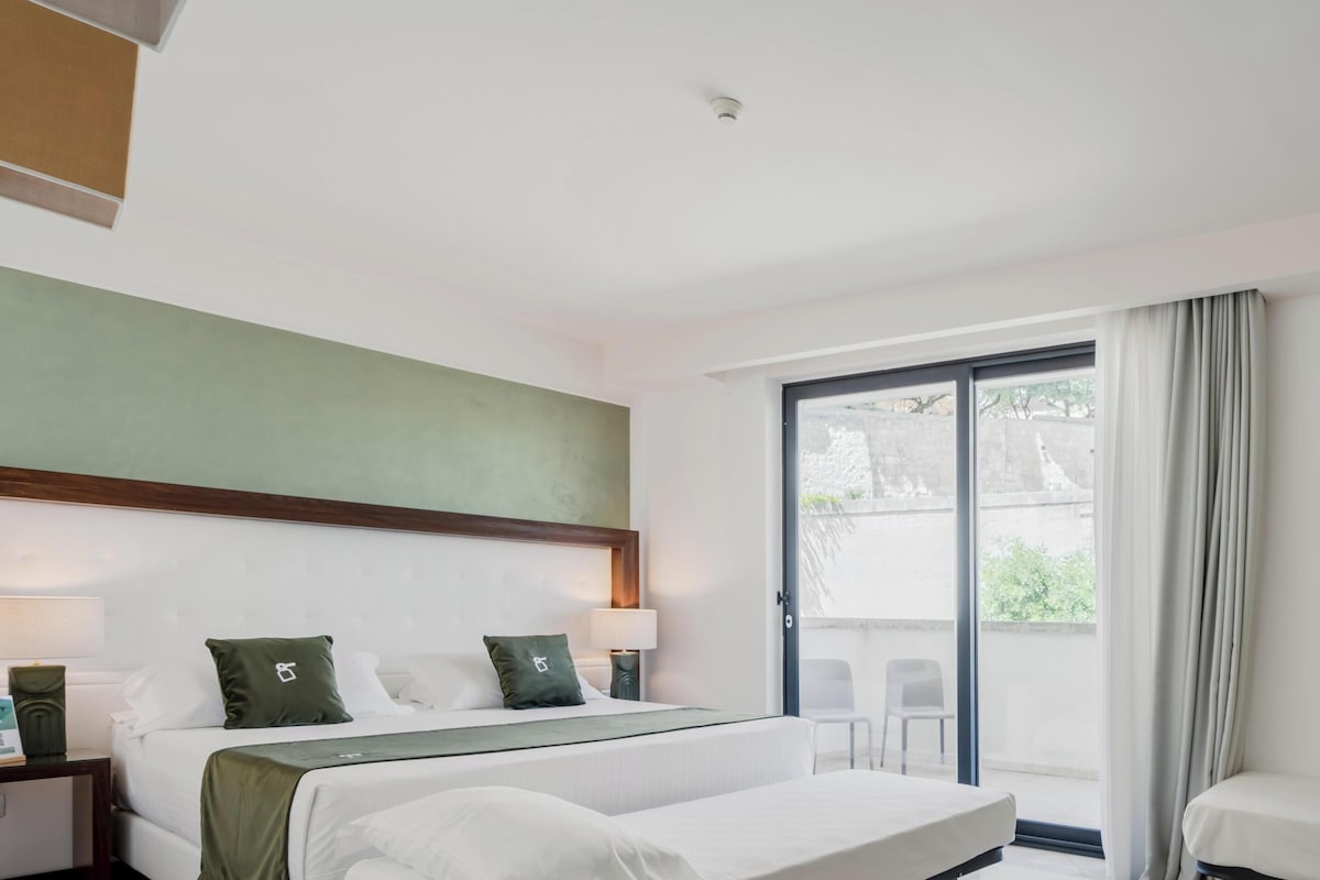 Habitación de hotel con cama, mesita de noche, lámpara y vistas al exterior a través de una puerta corrediza.