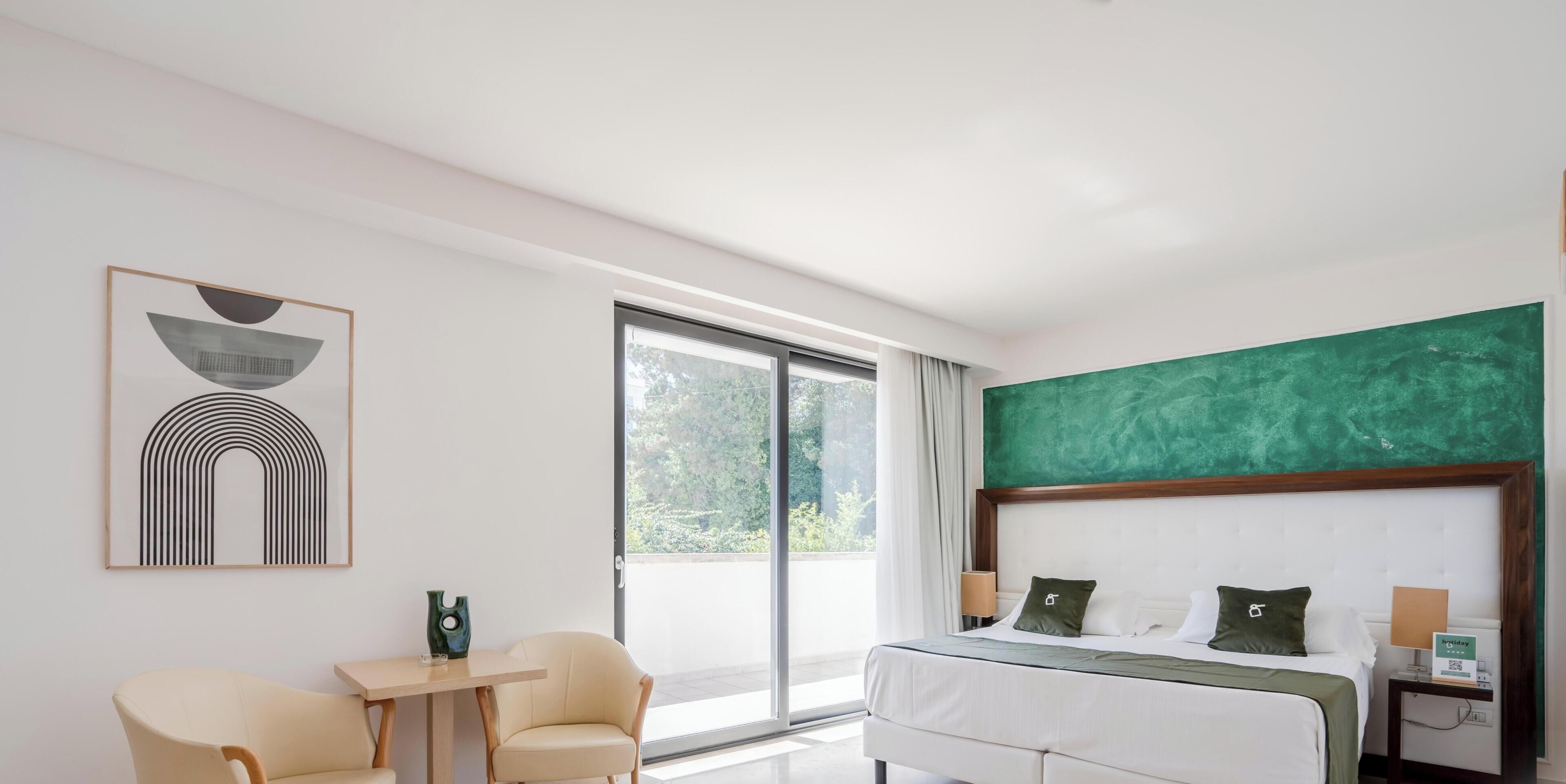A modern hotel room with a bed, two chairs, a table, a large window, and artwork on the wall.