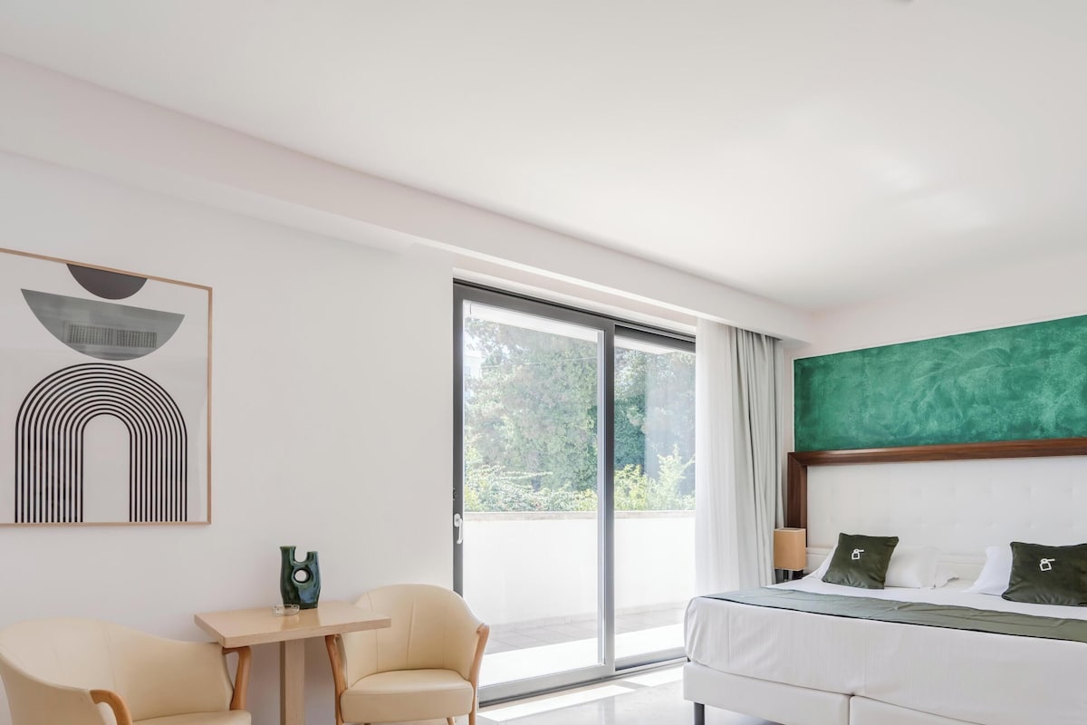 Una habitación de hotel moderna con una cama, un escritorio, dos sillas, un ventanal grande y una pared de acento verde.