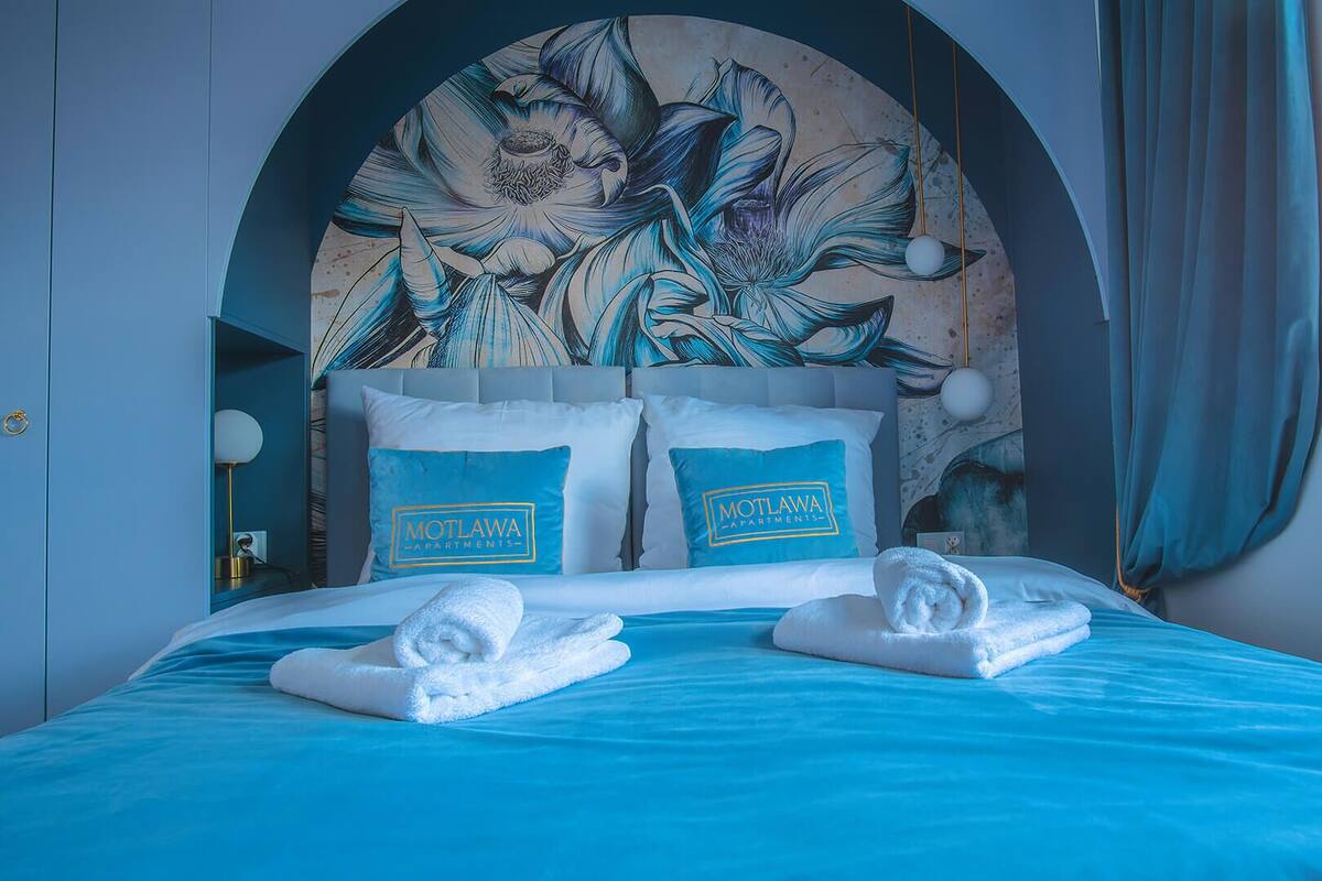 A hotel room with a large floral mural on the headboard, two pillows with the hotel name, a neatly made bed, and a blue bedspread.