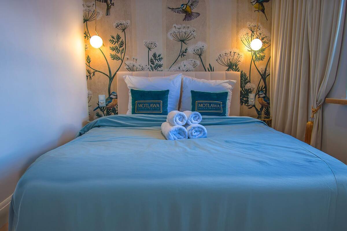 A hotel room with a bed, two pillows with 'Motlawa Apartments' branding, a bedside table, and wall art depicting birds and flowers.