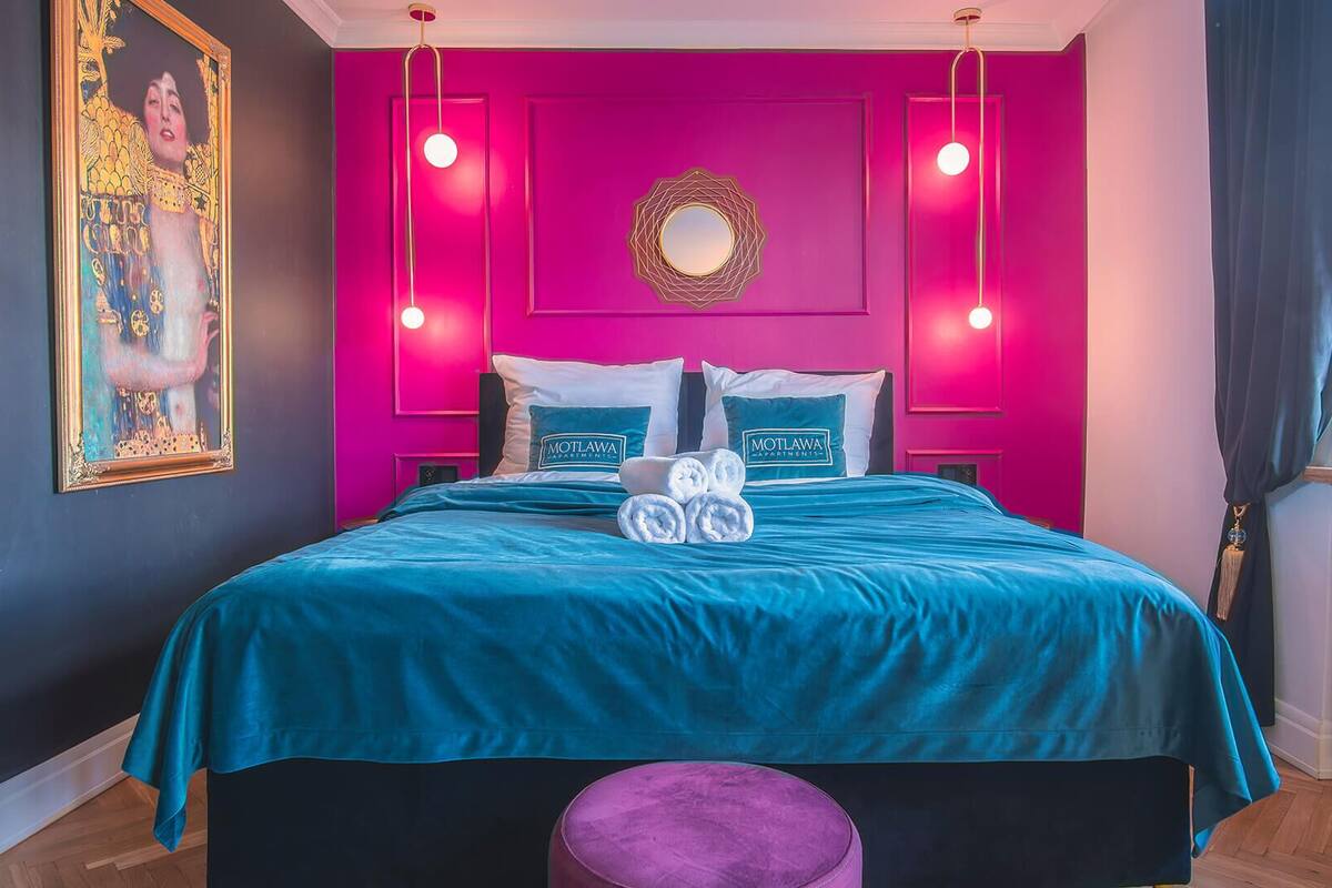 A bedroom with a teal bedspread, a purple ottoman, and a painting on the wall.