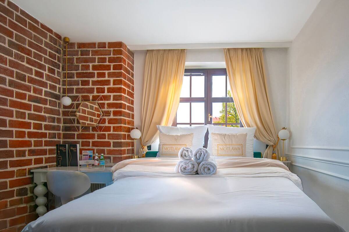 A hotel room with a brick wall, a large bed with white bedding, two bedside tables with lamps, and a window with curtains.