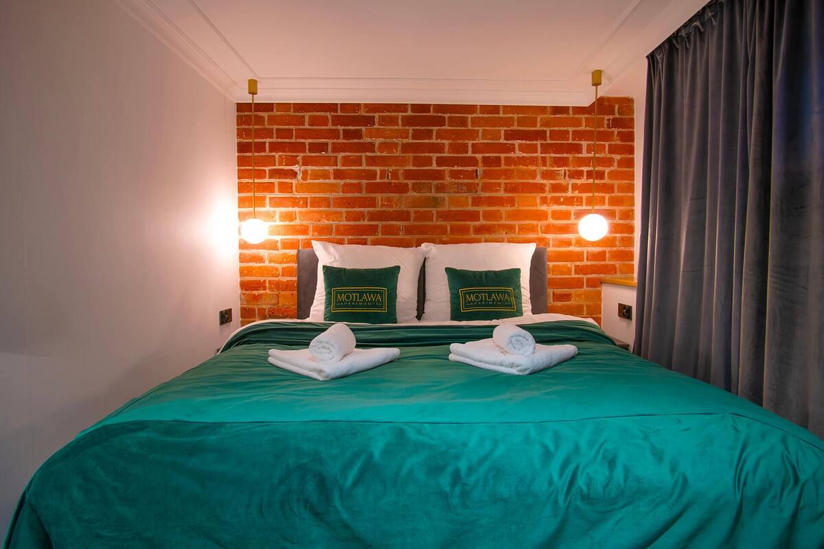 A hotel room with a brick wall, two Motlawa branded pillows, a green bedspread, and a pair of towels on the bed.