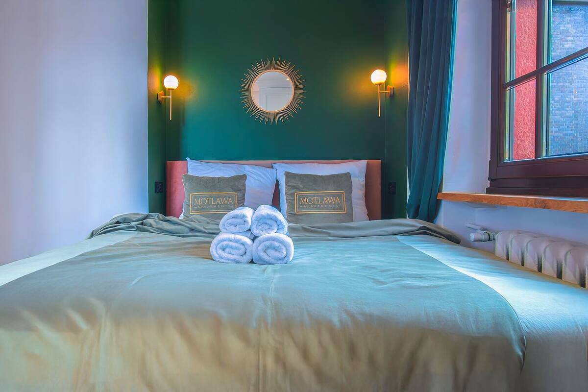 A neatly made bed with rolled towels, two wall sconces, a mirror, and a window with curtains.