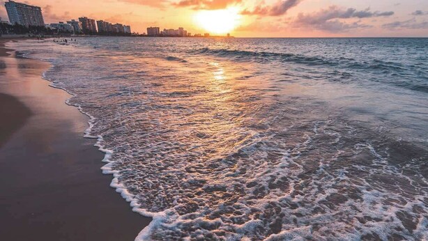 A beach with waves at sunset, buildings in the background, and a clear sky. - Steps from Beach 2 Bed 2 Full Bath w/PK  @ ESJ (Carolina)