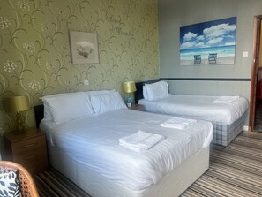 A hotel room with two beds, a painting of a beach scene, and a floral wallpaper pattern. - The Holmlea  (Blackpool)