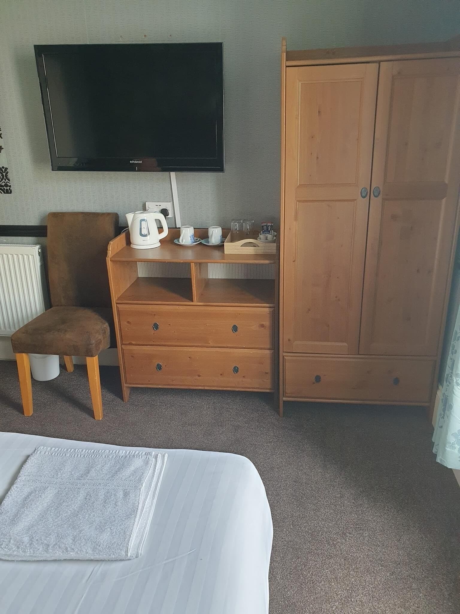 A hotel room with a bed, a wooden wardrobe, a television mounted on the wall, a small table with a kettle and cups, and a chair.