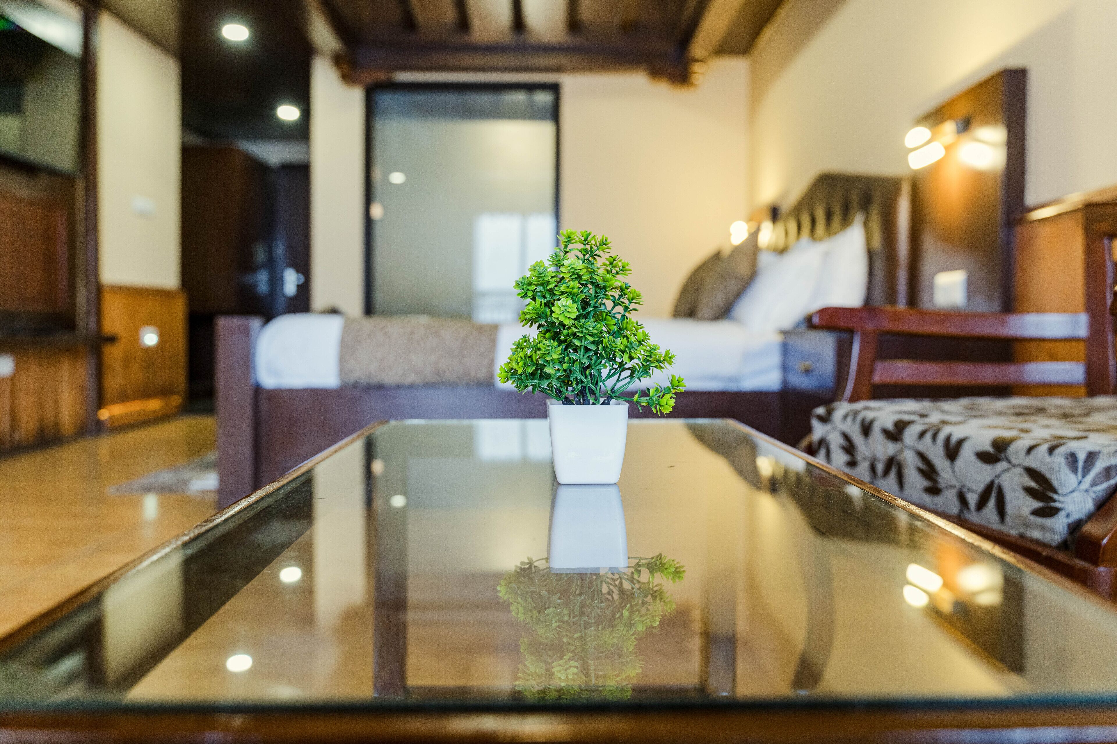 A modern hotel room with a glass-top table, a small potted plant, a bed with a patterned bedspread, and a wooden chair.