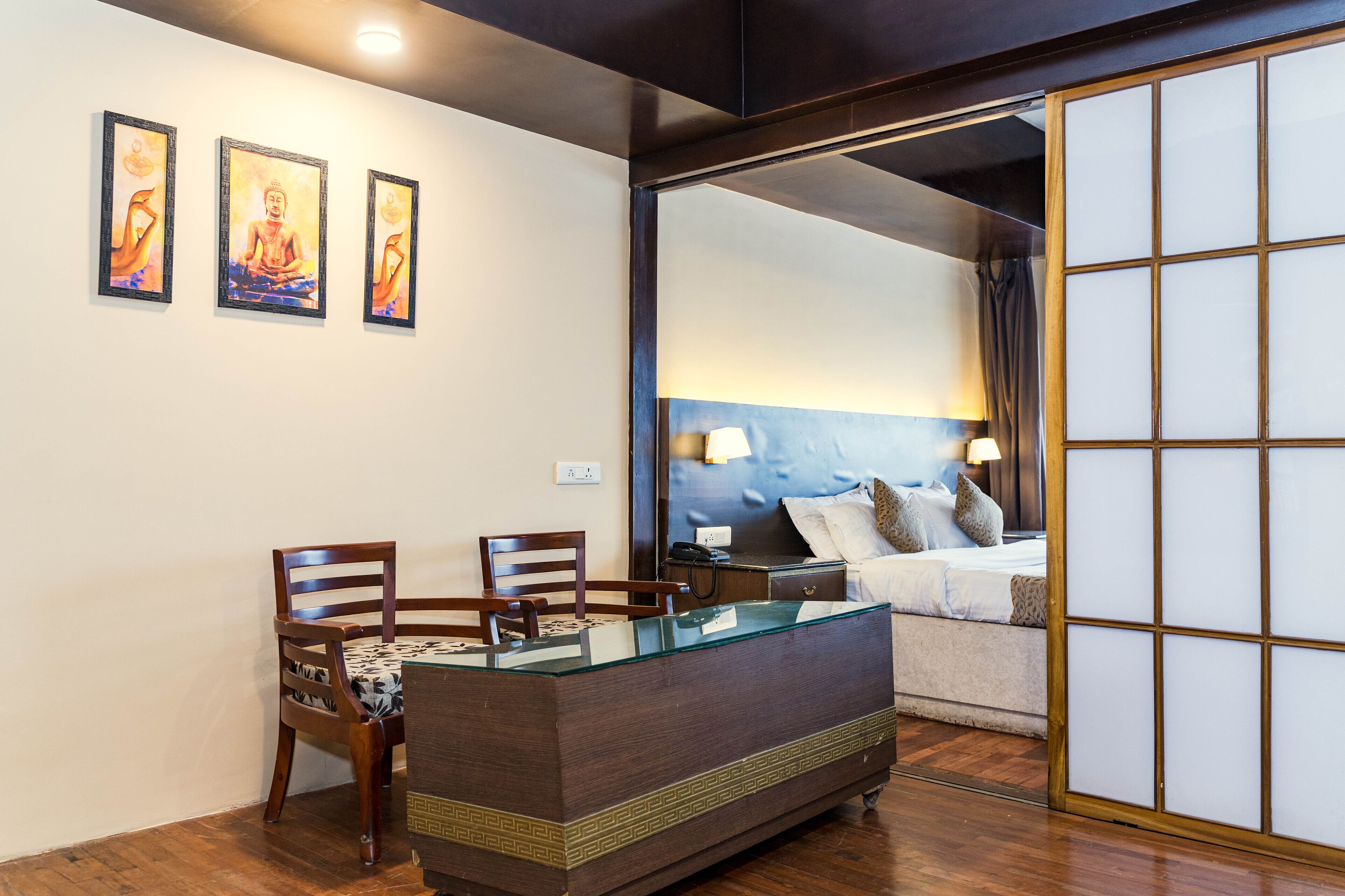 A room with a wooden desk, two chairs, a bed with a blue headboard, and three framed pictures on the wall.
