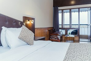 A hotel room with a large bed, a wooden headboard, a desk with a chair, and a view of the balcony. - Bliss Hungry Jack (Gangtok)