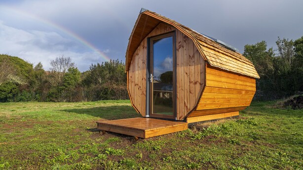A wooden cabin with a large sliding door and a small porch, set in a grassy field with a rainbow in the background.