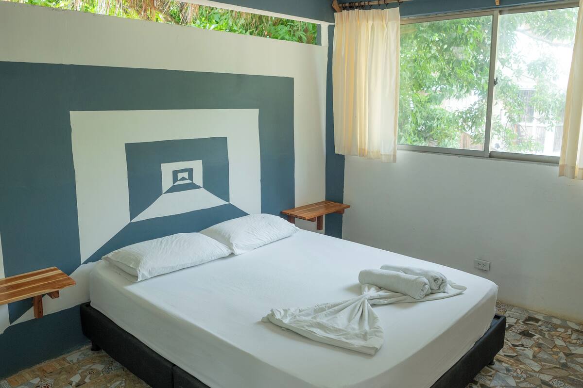 Una cama bien tendida con ropa y toallas blancas, una mesita de noche de madera, una pequeña mesa de madera y una ventana con cortinas.