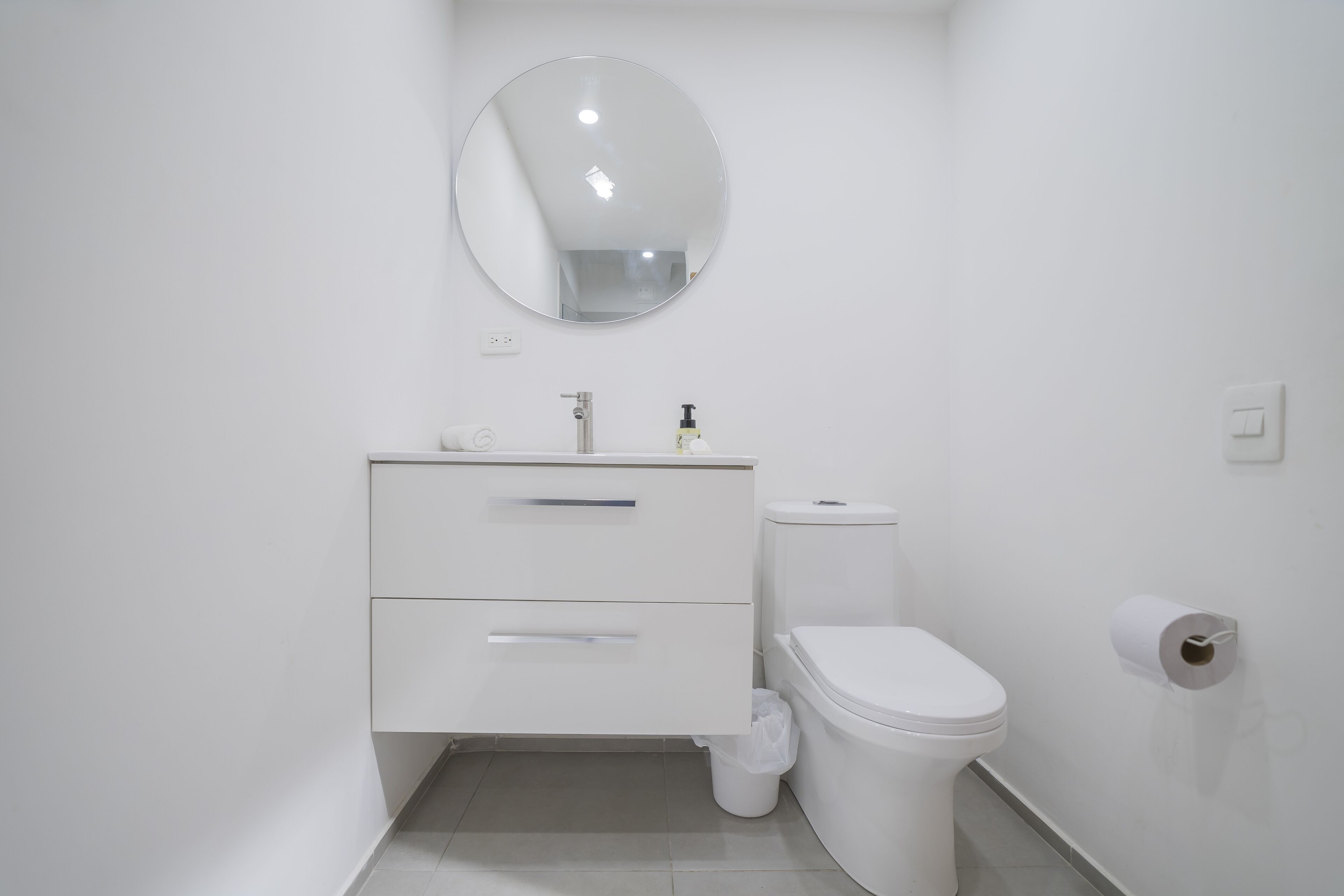 A white bathroom with a round mirror, a white toilet, and a white sink with two drawers.