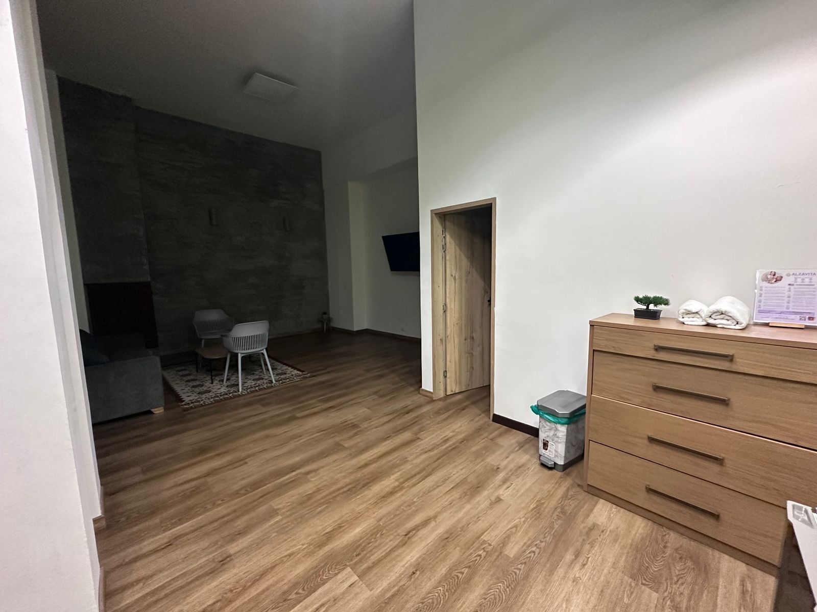 A room with a wooden floor, a small wooden cabinet, a trash bin, and a door leading to another room.