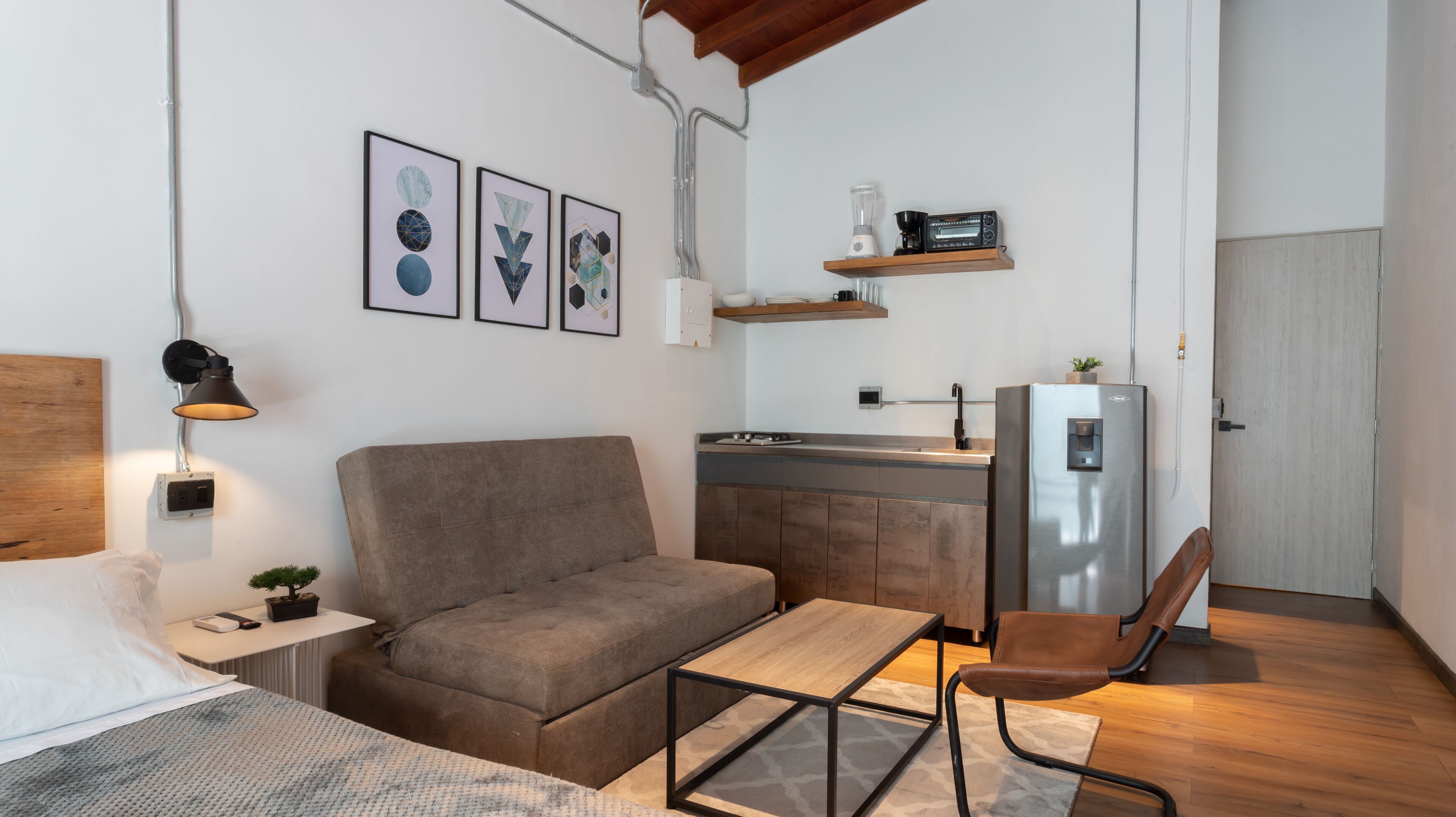 A compact living space with a sofa, a small table, a chair, a kitchenette, and a bed.