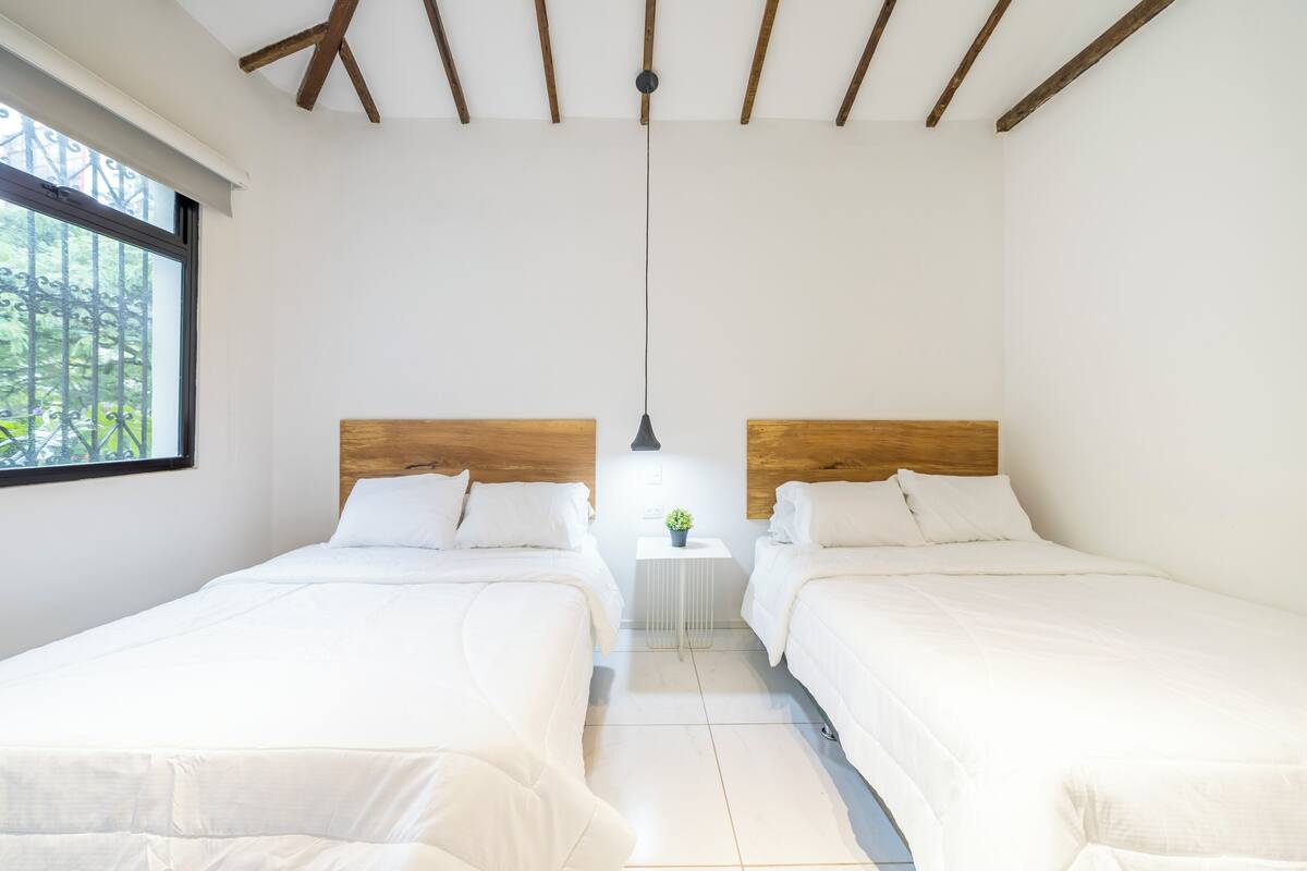 A room with two beds, a small white table with a plant, and a window with a view of greenery.