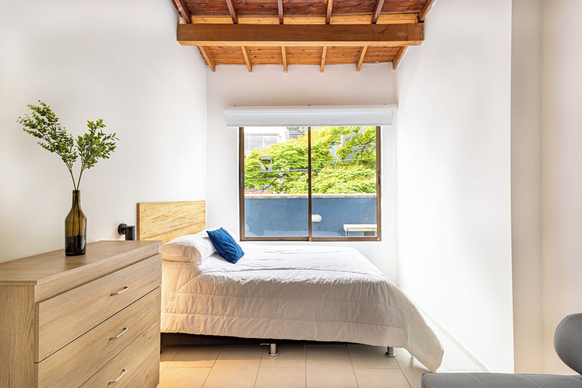 A bedroom with a bed, a dresser, a window with a view of greenery, and a wooden ceiling.