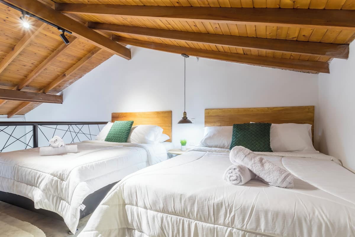 A room with two beds, wooden beams, a small table with a plant, and a balcony with a railing.
