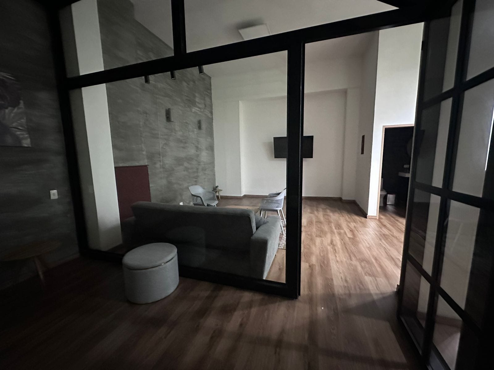 Sala de estar moderna com um sofá cinzento, um banquinho redondo pequeno e chão de madeira. Há uma grande porta de vidro que dá para outro cômodo com uma televisão e uma poltrona.